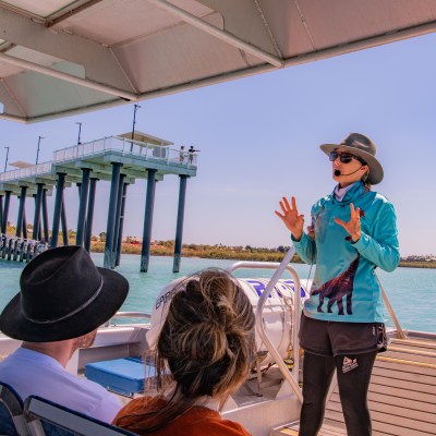 Guide speaking to tourists on a boat near a long pier over turquoise water.