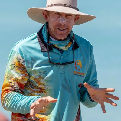 Man in a wide-brim hat, wearing a colorful shirt, gestures with hands against a blue sky background.
