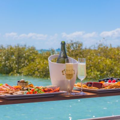 Floating table with assorted food, champagne, and glasses over a sunny waterfront view.