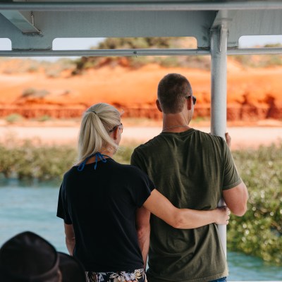 Couple standing on a boat, embracing, with a view of red cliffs and water.