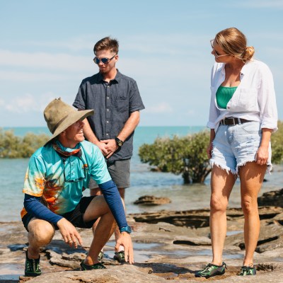 Three people on a rocky shoreline, with one crouching and two standing, ocean and trees in the background.