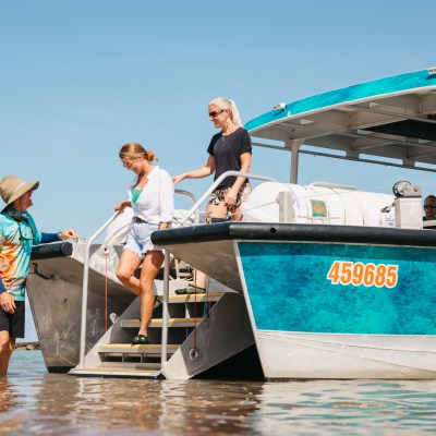 Two women descending stairs from a tour boat to shallow water, assisted by a man in a hat.