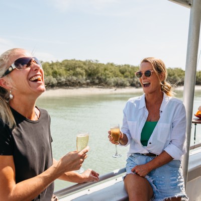 Two women laughing and holding drinks on a boat near a riverbank.