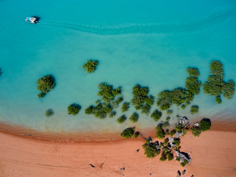 Aerial view of a boat on turquoise water near a sandy beach with scattered lush green vegetation.