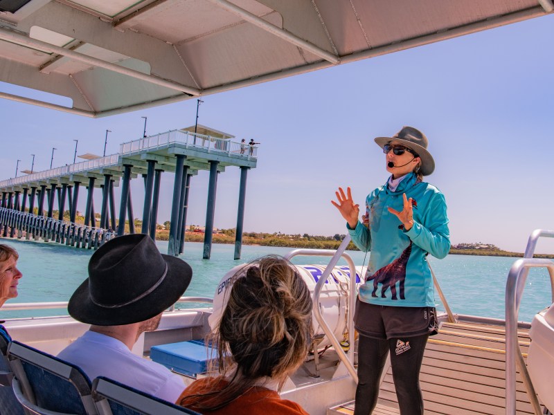 Tour guide speaking to passengers on a boat near a long pier over turquoise water.