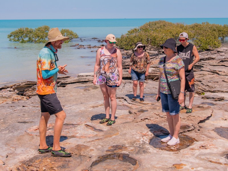 A group of people explore rocky terrain by the sea on a sunny day, with clear blue water in the background.