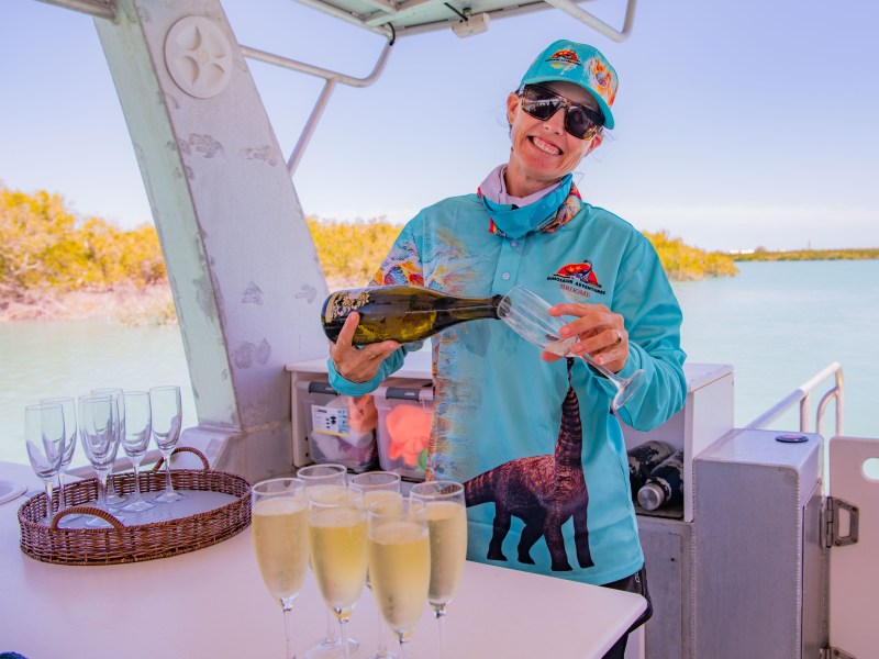 Person pouring champagne on a boat with water and trees in the background.