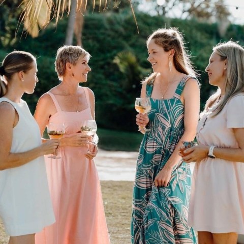 Four women in summer dresses chat and hold drinks outdoors near palm trees.