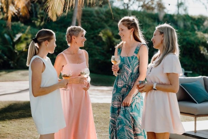 Four women in summer dresses chat and hold drinks outdoors near palm trees.