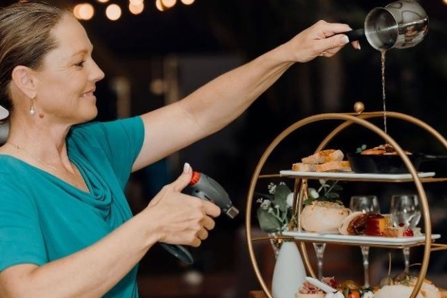 Smiling woman in flower hairpin pouring liquid over tiered food display while holding a kitchen torch.