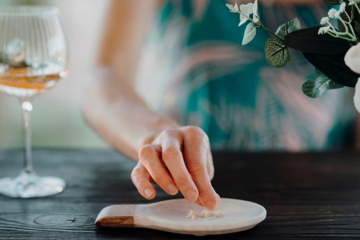 Hand reaching for salt on a wooden board, next to flowers and a wine glass.