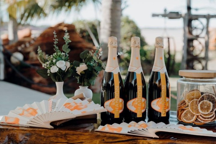 Three bottles of champagne, paper fans, and a jar of dried citrus on a wooden table with flowers.