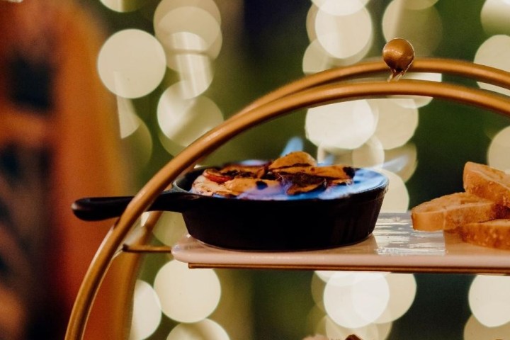 Small skillet with food on flaming burner, bread slices on tray, bokeh lights background.