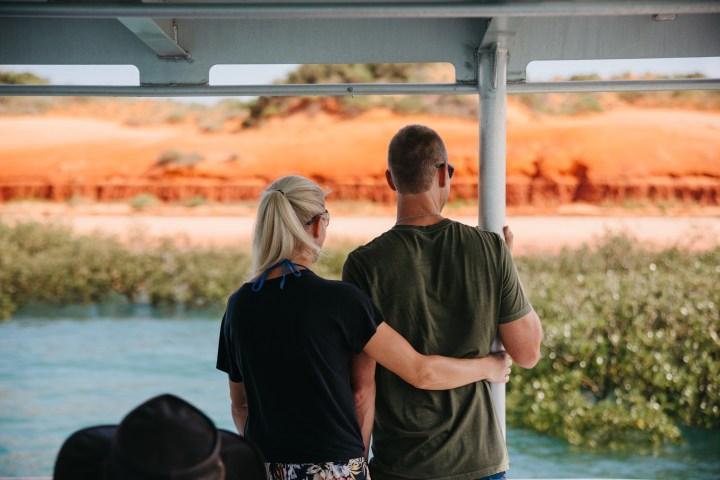Couple embracing on a boat, looking at red cliffs and water in the background.