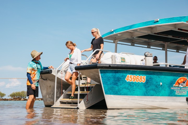 Two people disembark a blue boat onto a sandy shore, assisted by a person in colorful attire.