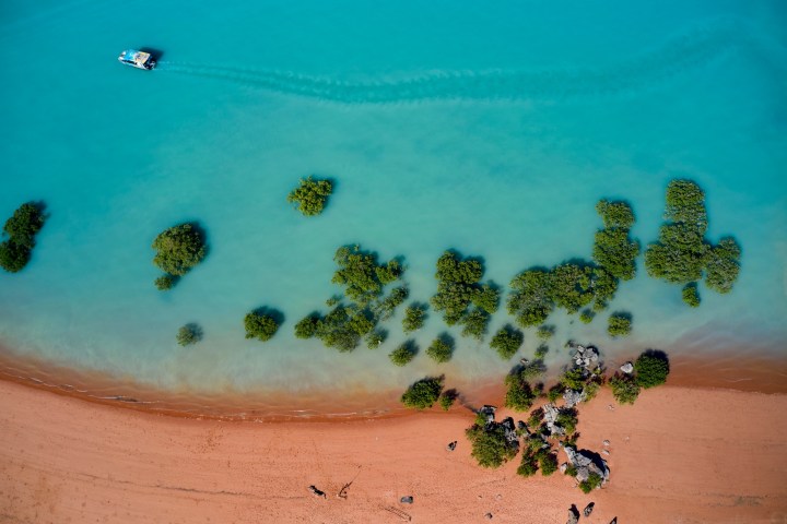 Aerial view of a boat on turquoise water near a sandy shore with green plants.
