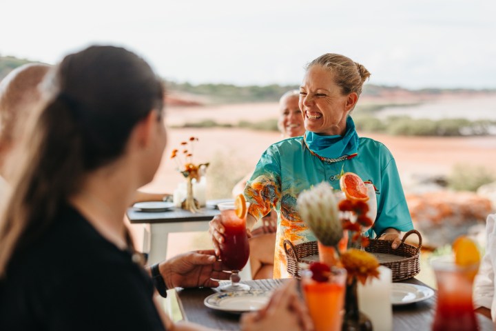 Smiling woman serves drinks to people at an outdoor table by the beach.