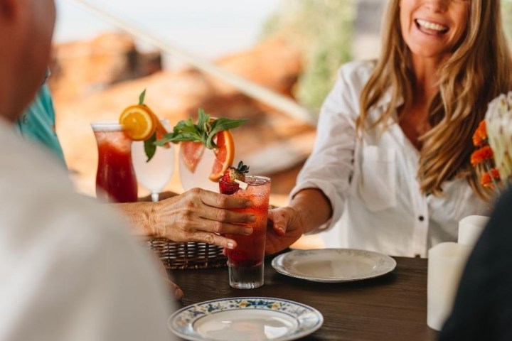 Smiling woman at table with colorful drinks and plates, sharing a drink.
