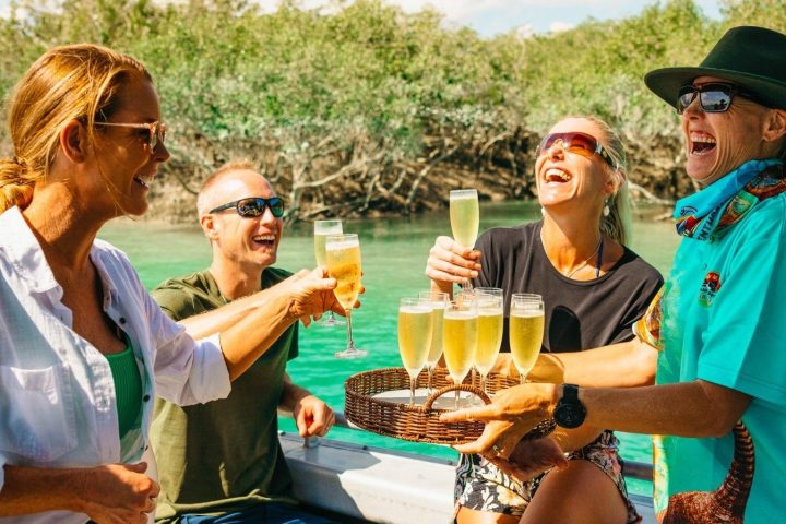 Four people on a boat laughing and holding glasses of champagne by a green river.