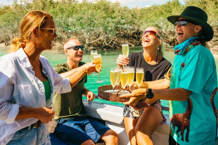 Four people on a boat laughing and holding glasses of champagne by a green river.
