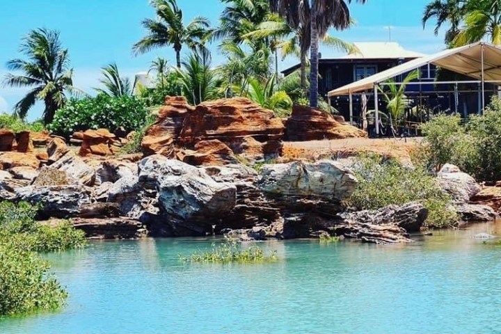 Tropical scene with rocky shore, turquoise water, and palm trees near a building.