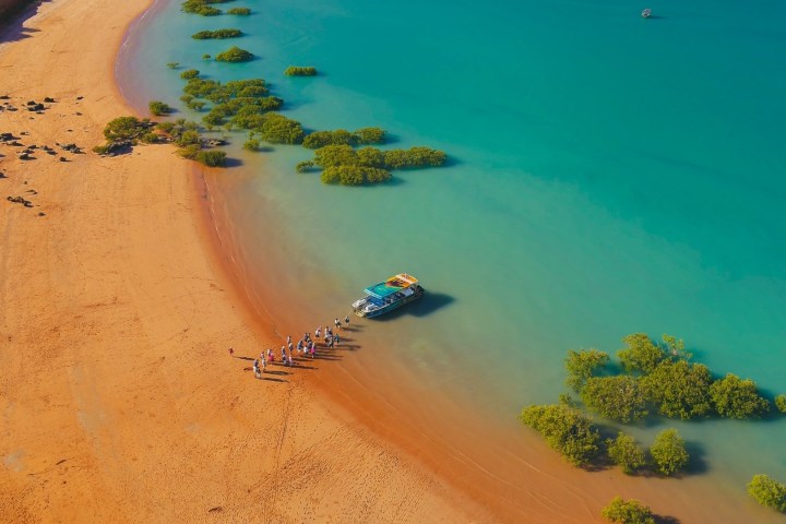 Aerial view of a beach with a boat and people by the shoreline and scattered green bushes in clear blue water.