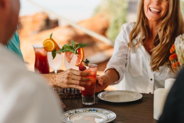 Smiling woman handing a strawberry drink to a person at a table with colorful beverages.