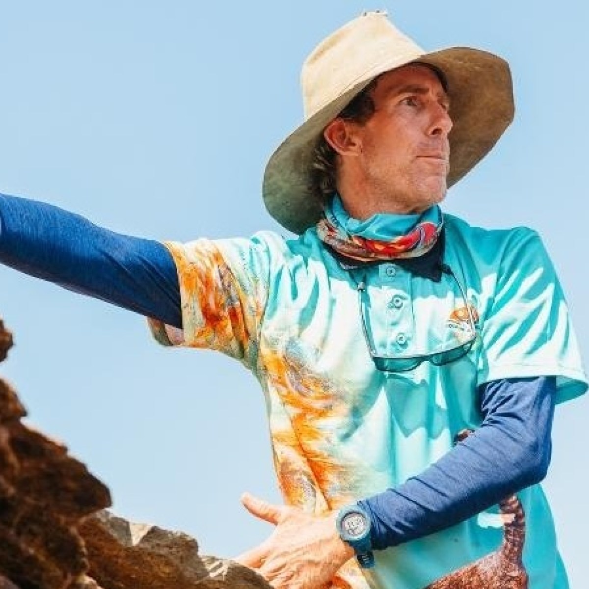 Person in hat and colorful shirt gestures outdoors amid rocky terrain.