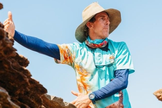 Person in hat and colorful shirt gestures outdoors amid rocky terrain.
