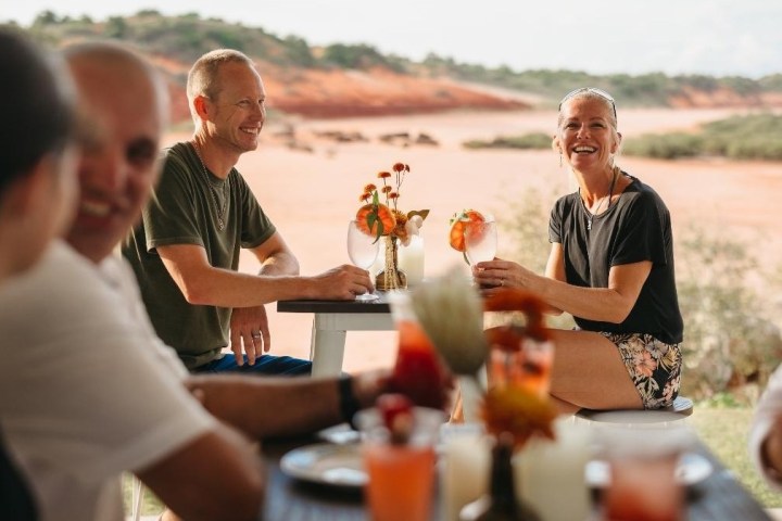 Group of people smiling, enjoying drinks at an outdoor table with a scenic background.