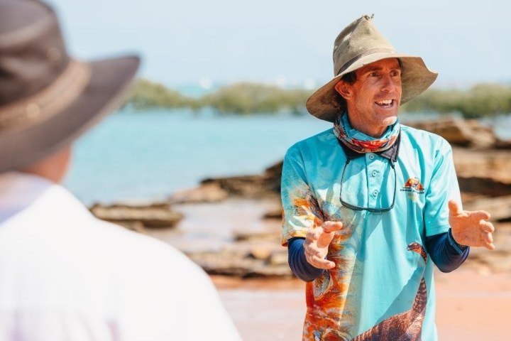 Man in hat talks passionately on a rocky beach with water and vegetation in the background.