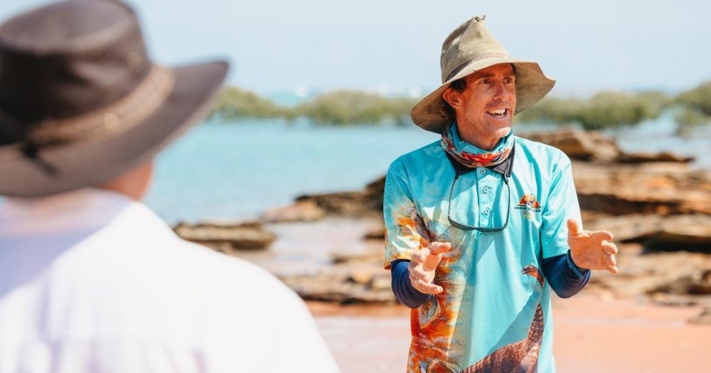 Man in hat talks passionately on a rocky beach with water and vegetation in the background.
