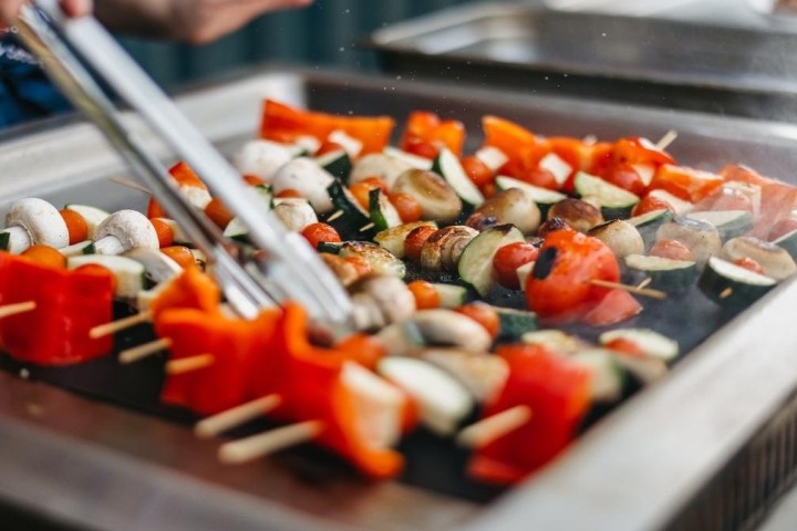 Vegetable skewers with mushrooms, peppers, and zucchini on a grill being turned with tongs.