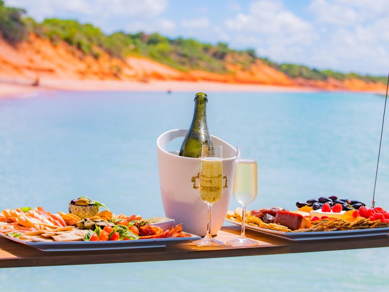 Floating table with snacks, champagne, and glasses over water with cliffs in background.