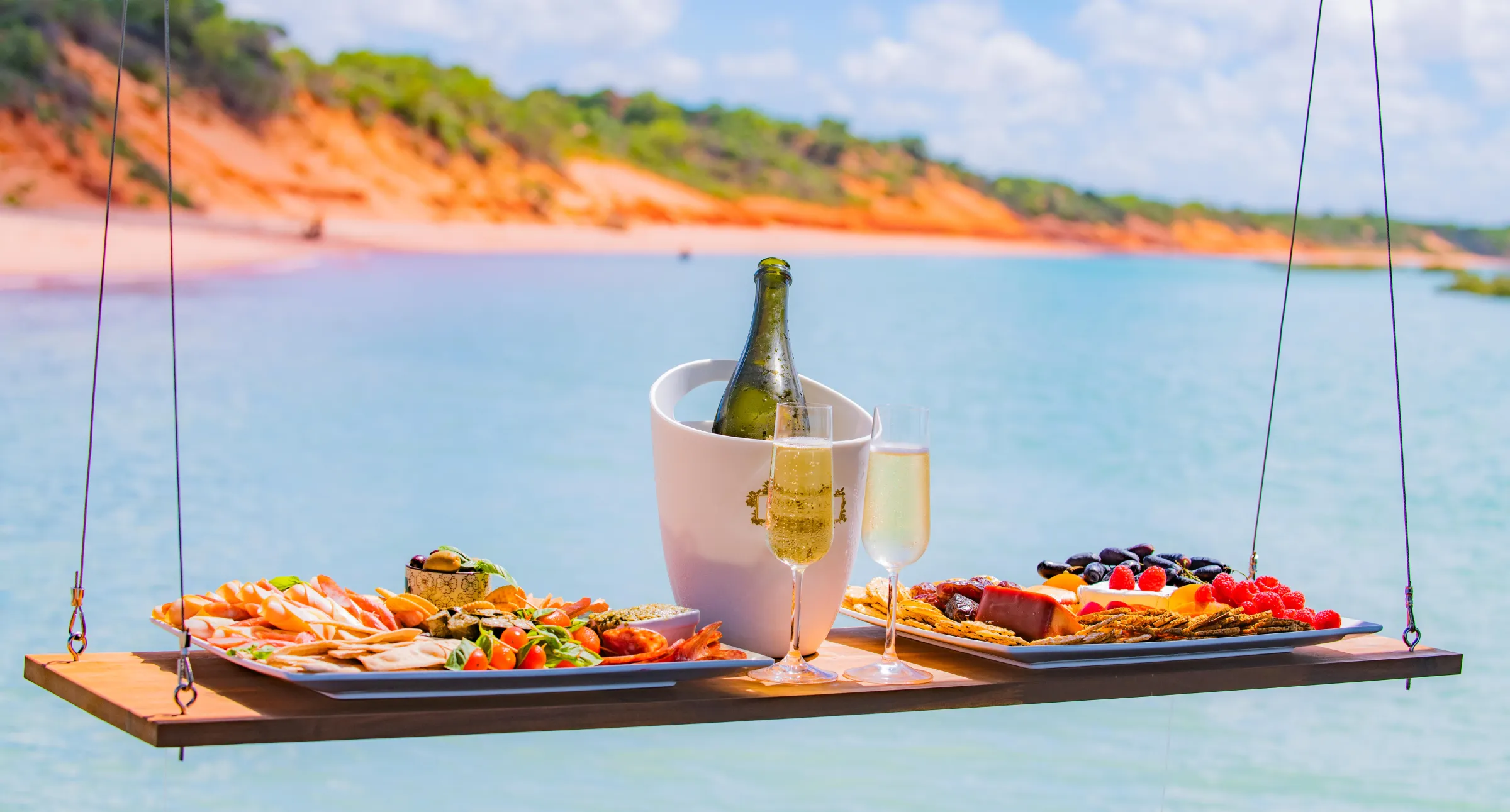 Floating table with snacks, champagne, and glasses over water with cliffs in background.