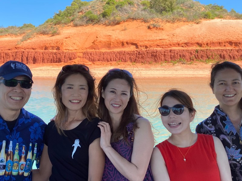 Five people smiling in front of red cliffs with clear blue water and sky.