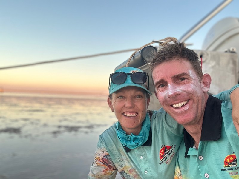 Two people smiling on a beach at sunset, wearing turquoise shirts and hats.