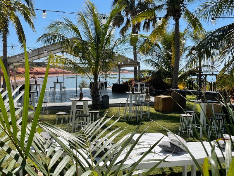 Outdoor beachside cafe with palm trees, tables, chairs, and string lights under a clear blue sky.
