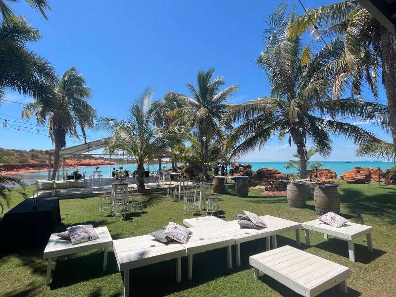 Seaside resort with palm trees, white benches, and tables overlooking the ocean under a clear blue sky.