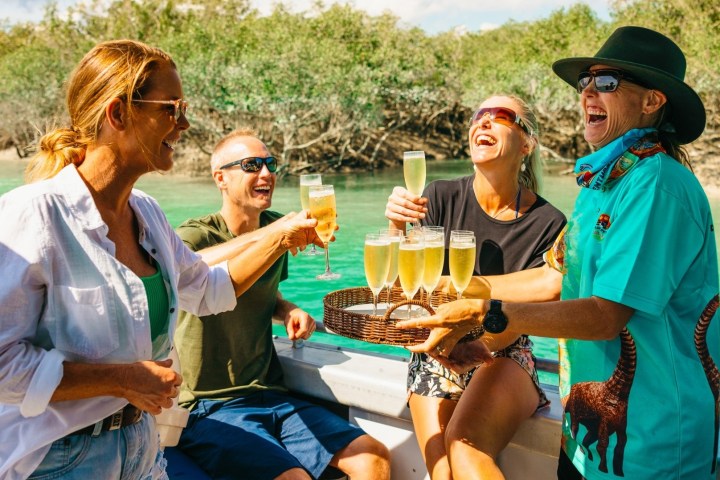 Group of four people laughing and holding drinks on a boat near a mangrove area.