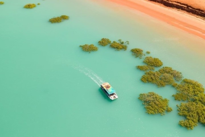 Aerial view of boat in turquoise water near green foliage and orange sandy shoreline.
