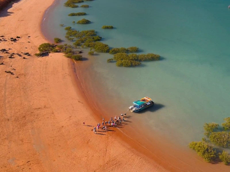 Aerial view of people near a beached boat on a sandy shore with green patches in turquoise water.