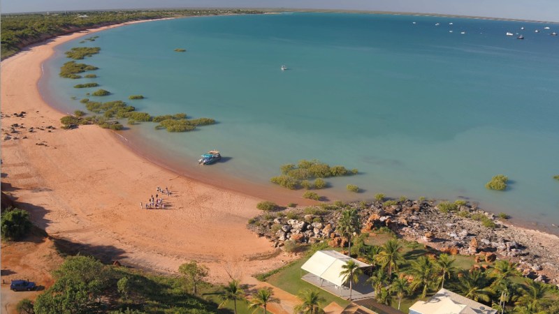 Aerial view of a sandy beach with people, a boat, and scattered vegetation along a turquoise bay.