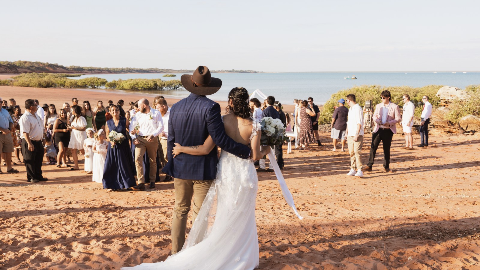 Bride and groom facing guests on a sandy beach with ocean in the background.