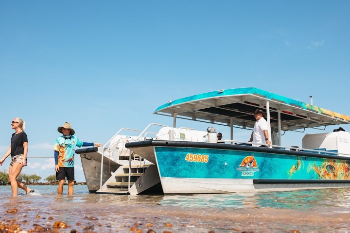 People walking on shallow water near a docked blue tour boat under a clear sky.
