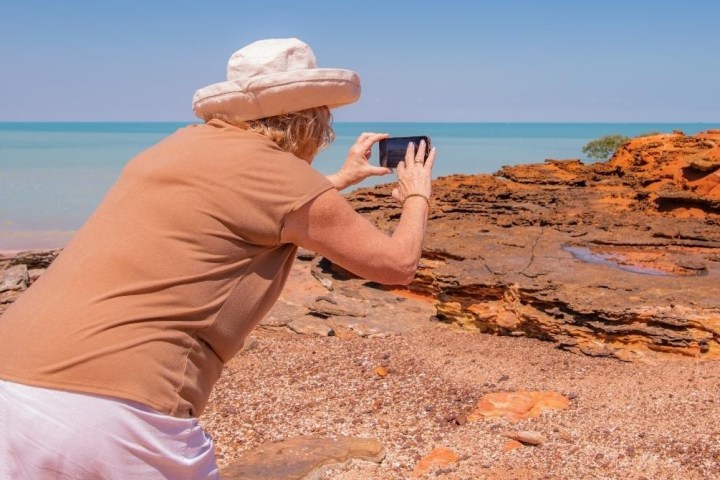 Person in a sun hat taking a photo of rocky beach with clear blue sea.