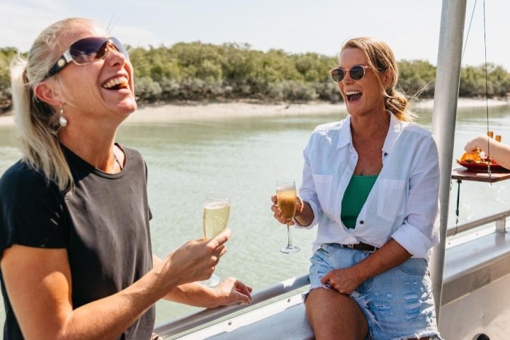 Two women laughing and holding drinks on a boat by a river.