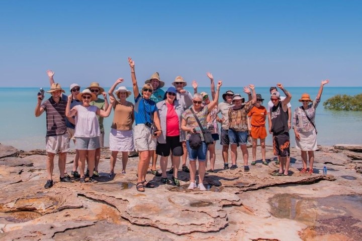 Group of people waving on rocky beach with blue sky and ocean in background.