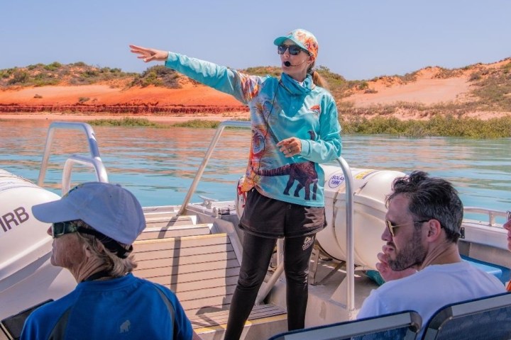 Guide pointing while talking to tourists on a boat with scenic red cliffs in the background.