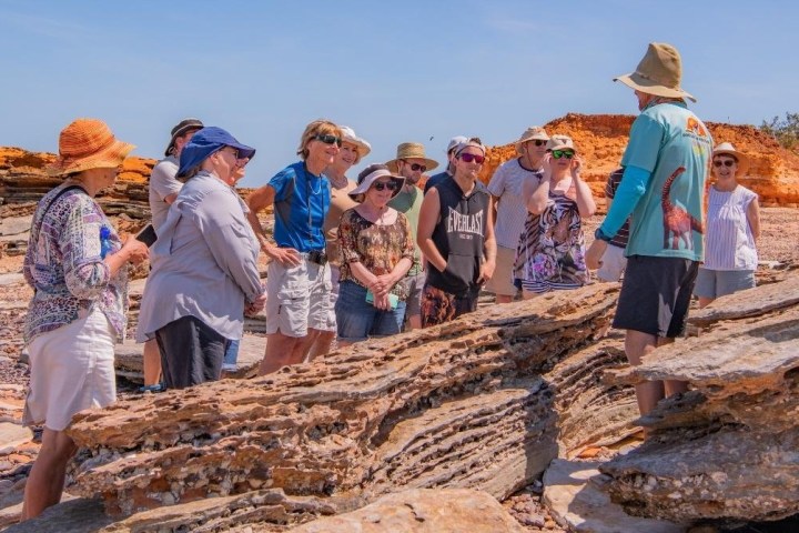 Group of people on rocky cliff under clear sky, listening to a tour guide.